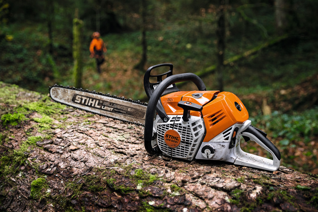 Orange STIHL chainsaw resting on a mossy log in a forest, with a person in orange gear blurred in the background.