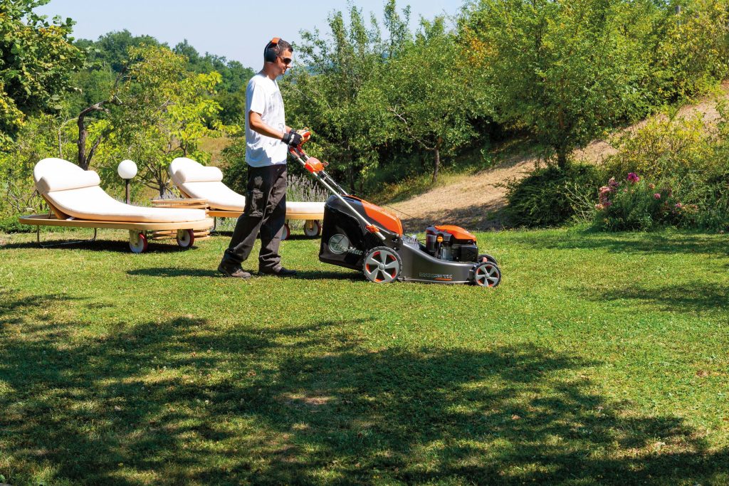 Man wearing ear protection pushes an orange and black lawn mower across a sunny backyard with white lounge chairs and green trees nearby.