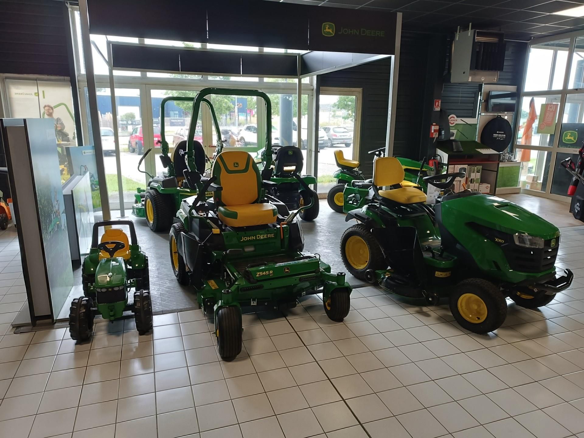 Showroom filled with green and yellow John Deere riding lawn mowers and small tractors on a tiled floor.]
