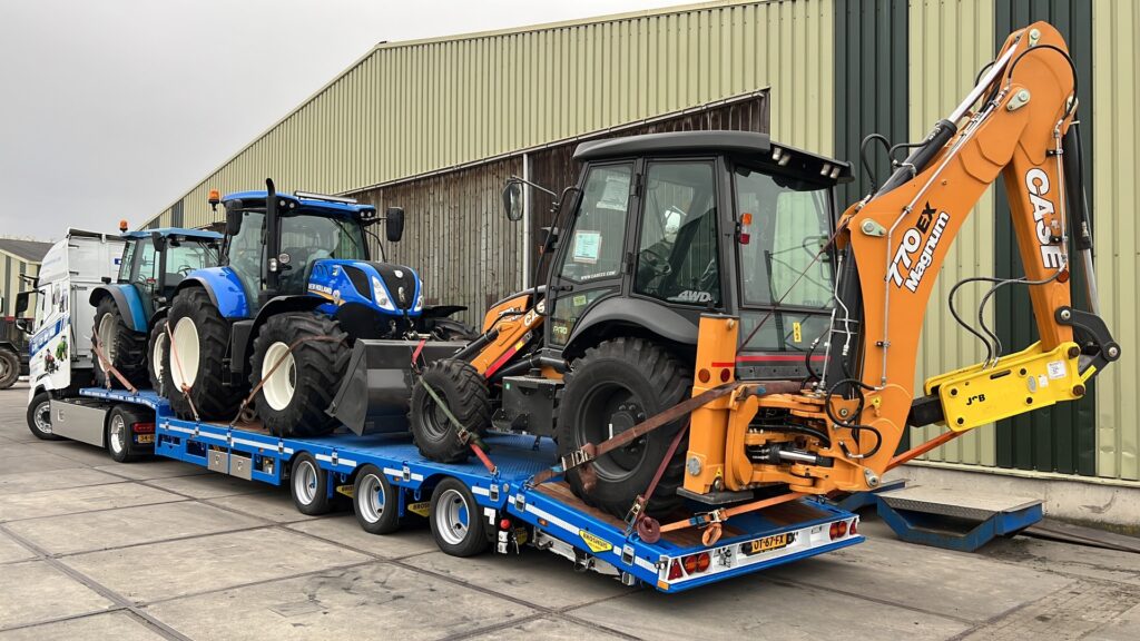 Flatbed trailer hauling three tractors: two blue tractors and an orange Case backhoe loader at the rear, outside a warehousework area.