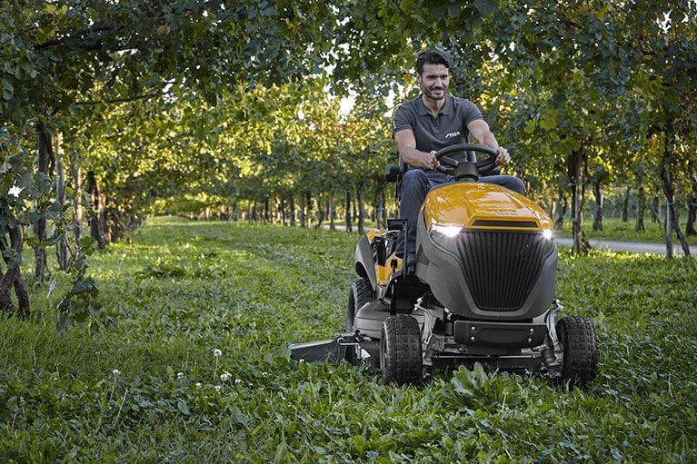 Man operating a yellow riding lawn mower through a sunlit orchard.